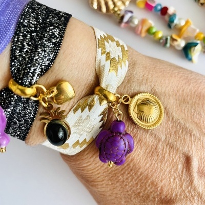 Pulseiras coloridas com pendentes dourados numa mão, fundo branco com acessórios coloridos