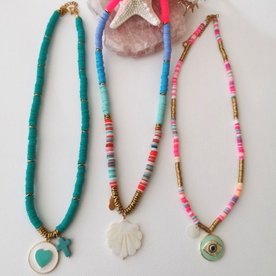 Três colares coloridos com pendentes e conchas decorativas ao fundo