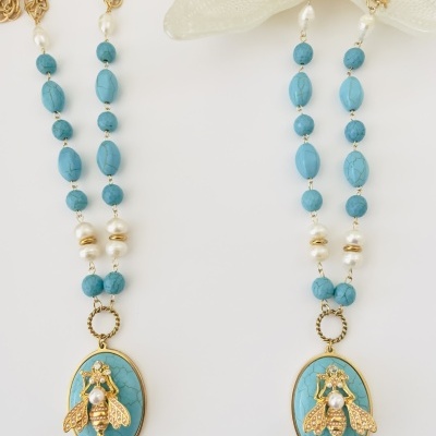 Colar dourado com contas azuis e pendente de abelha dourada sobre base azul-turquesa