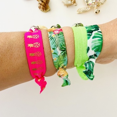 Três pulseiras de tecido elástico com padrões tropicais e pinhas douradas no pulso, sobre fundo branco.