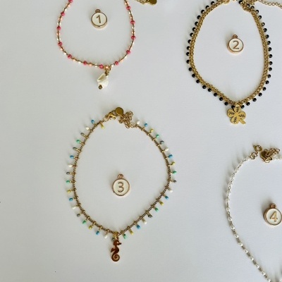 Quatro pulseiras douradas com contas coloridas e pérolas, cada uma com pendente distinto, dispostas em fundo branco com etiquetas numeradas e estrela do mar