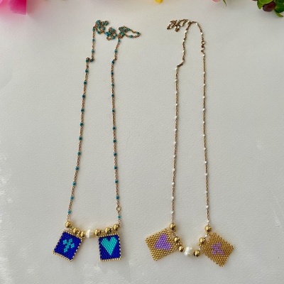 Dois colares com pingentes de contas coloridas e pérolas sobre superfície branca com flores ao fundo.