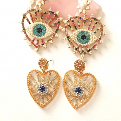 Dois pares de brincos coloridos, um em formato de olhos com pérolas e pedras coloridas e outro em formato de coração dourado com pedras azuis e brancas