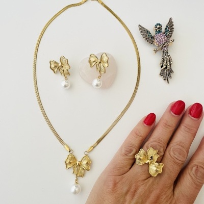 Conjunto de joias douradas com laços e pérolas numa superfície branca ao lado de broche colorido e mão com unhas vermelhas.