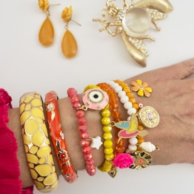 Pulso com várias pulseiras coloridas, brincos amarelos pendentes e broche dourado em superfície branca