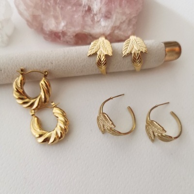 Três pares de brincos dourados com diferentes texturas e padrões em fundo branco, pedra rosa e suporte cilíndrico bege