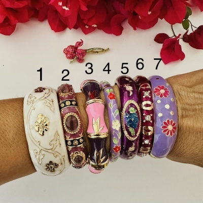 Sete pulseiras coloridas com padrões florais e detalhes dourados, numeradas de 1 a 7, sobre pele com flores vermelhas em fundo branco.
