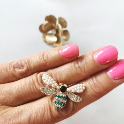 Anel dourado em forma de abelha com pérolas e pedras coloridas numa mão com unhas rosa.
