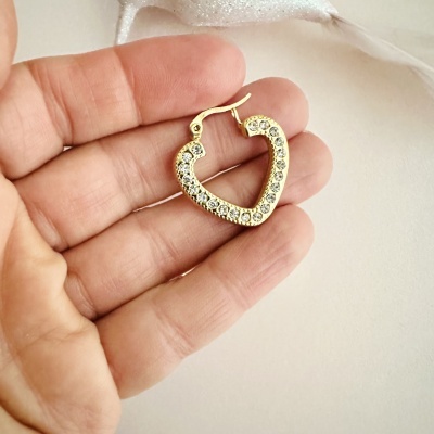 Brinco de coração dourado com pedras brilhantes numa mão sobre fundo branco