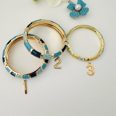 Três pulseiras douradas com detalhes azuis e verdes e pendentes numerados, sobre fundo branco com flor azul