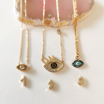 Colares dourados com pendentes de olhos estilizados e pingentes numéricos sobre superfície branca com cristal rosa