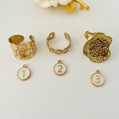 Três anéis dourados com medalhas numeradas 1, 2 e 3 sobre superfície branca, com flor decorativa ao fundo