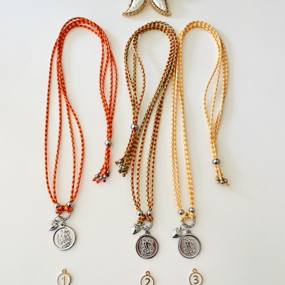 Três colares trançados coloridos com pingentes metálicos e números 1, 2 e 3 sobre fundo branco com flor decorativa