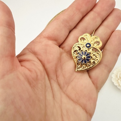 Colar dourado com pendente coração decorado azul numa mão com fundo branco