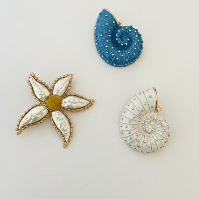 Três broches decorativos em metal dourado com pérolas falsas e esmalte azul, branco e amarelo em forma de concha e flor