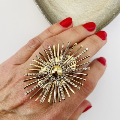 Anel dourado grande em forma de estrela com pedras brilhantes numa mão com unhas vermelhas