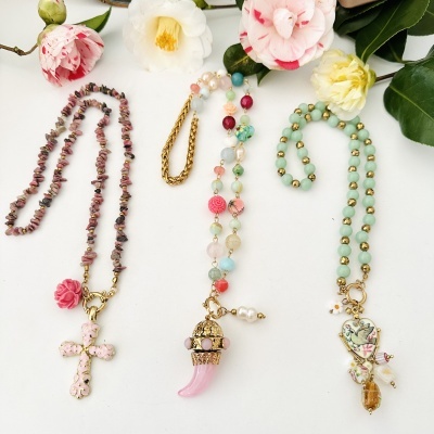 Três colares coloridos com pendentes, dispostos sobre fundo branco com flores ao redor.