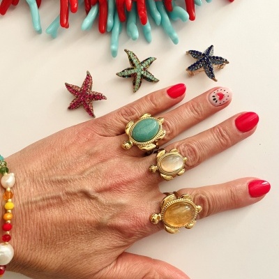 Mão com anéis de tartaruga dourados e pedras coloridas, pulseira e estrelas-do-mar coloridas ao fundo