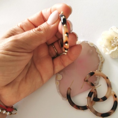 Brincos de argola castanhos e pretos com detalhes dourados numa mão, com geodo rosa e flor branca ao fundo