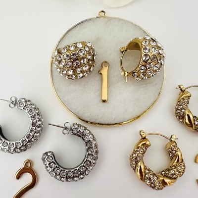 Brincos de argola dourados e prateados com pedras brilhantes sobre fundo branco com placa redonda de mármore.