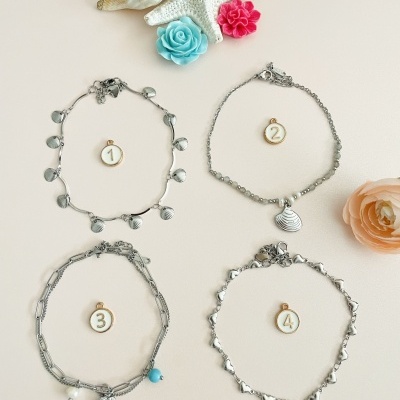 Quatro pulseiras prateadas decoradas com pendentes e contas sobre fundo claro com conchas e flores coloridas.