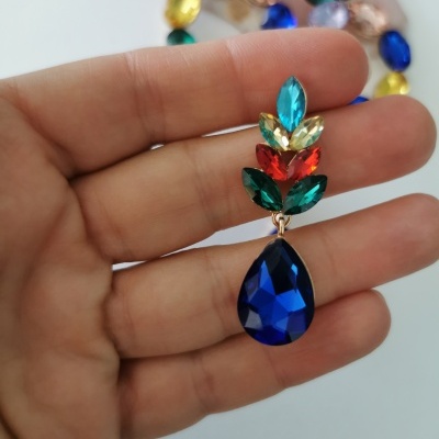 Brinco colorido com pedras facetadas em várias cores, pendurado numa mão sobre fundo branco