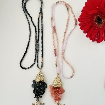 Dois colares com pendentes em forma de peixe, contas pretas e rosa, e flor vermelha ao lado.