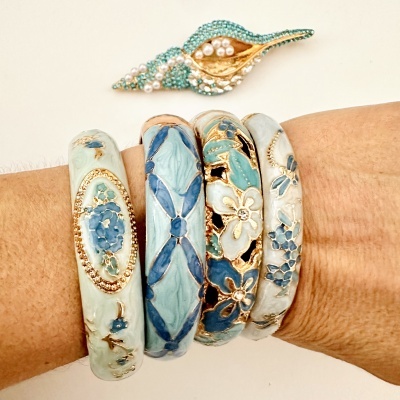 Pulseiras esmaltadas em azul e branco com padrões florais e geométricos no pulso e broche azul com pérolas ao fundo