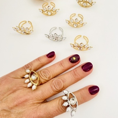 Anéis metalizados dourados e prateados com pedras brancas, alguns usados em mão com unhas pintadas vinho.