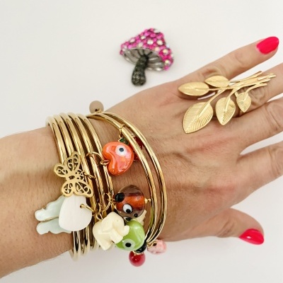 Mãos com pulseiras douradas e anéis com pingentes e design de folhas, fundo branco e broche de cogumelo rosa.