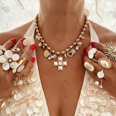 Colar e anéis dourados com pedras brancas e flores, mãos com unhas vermelhas, top branco com lantejoulas