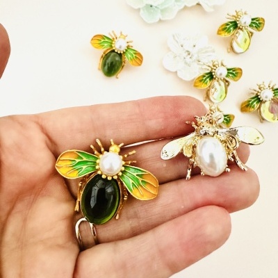 Broches de abelhas dourados com asas verdes e amarelas sobre mão e mesa branca