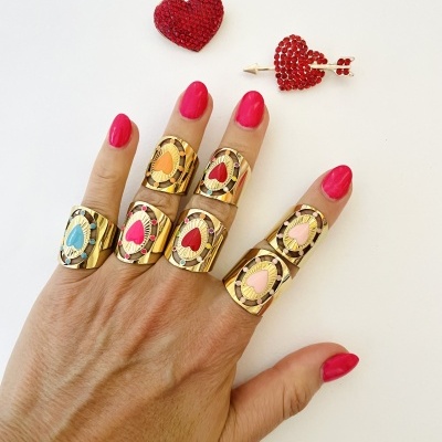 Mão com cinco anéis dourados com corações de várias cores e unhas pintadas de rosa