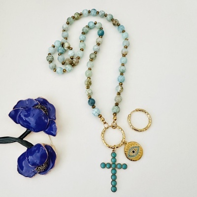 Colar com cruz turquesa, medalha dourada e argola sobre fundo branco e flores azuis.