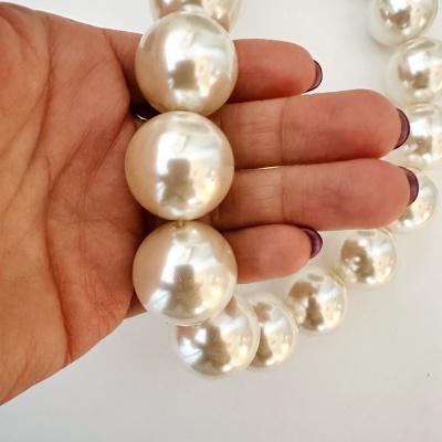 Colar de pérolas peroladas segurado por mão com unhas roxas em fundo branco