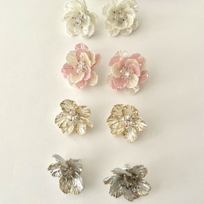 Cinco pares de brincos florais em várias cores dispostos em linha sobre fundo branco com flores naturais ao fundo.