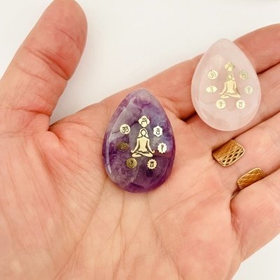 Dois pendentes em pedra roxa e rosa com símbolos dourados na mão de uma pessoa