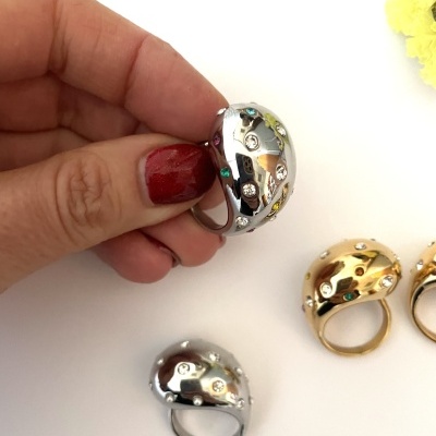 Anéis metálicos coloridos com pedras, prateados e dourados, em fundo branco com flores amarelas