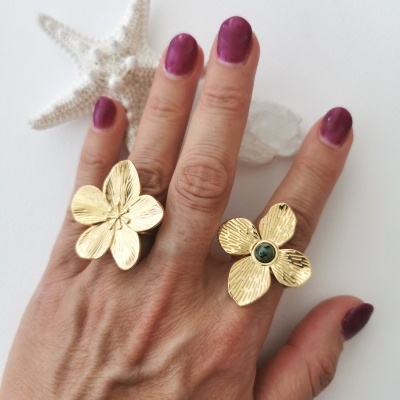 Mão com unhas roxas a usar dois anéis dourados em forma de flor, com estrela-do-mar e cristal ao fundo