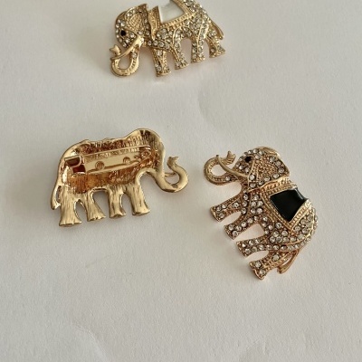 Três broches de elefantes dourados com pedras brilhantes e detalhes pretos sobre fundo branco.
