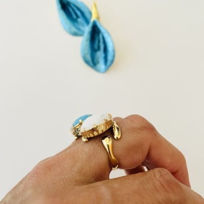 Anel dourado com pedra azul e branca numa mão com unhas pintadas, brincos azuis no fundo