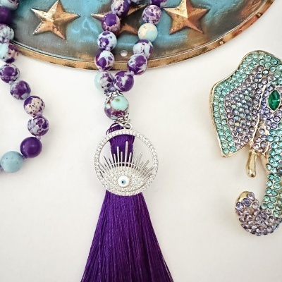 Colar com contas roxas, pendente de olho com cristais e franjas roxas ao lado de cavalo-marinho decorativo com strass.