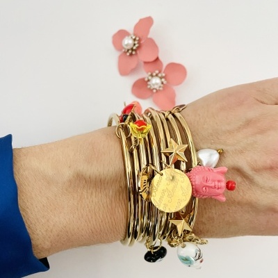 Mão vestindo várias pulseiras douradas com pendentes coloridos, sobre fundo branco com flores cor-de-rosa ao fundo.