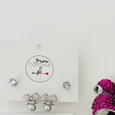 Brincos Maria Cotovia com pedras cristal e pérolas, cartão branco, ao lado de decoração floral rosa e preta