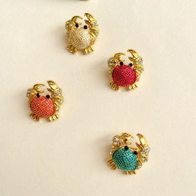 Broches de caranguejo coloridos com detalhes dourados e cristais, sobre fundo branco com concha verde.