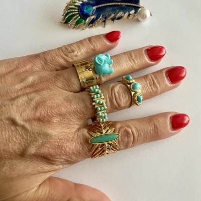 Mão com anéis dourados e coloridos e unhas vermelhas, com broche de pena azul e verde ao fundo