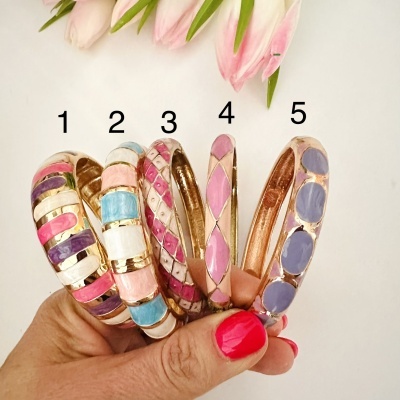 Cinco pulseiras coloridas de metal dourado com padrões esmaltados, seguradas por mão com unhas rosa.