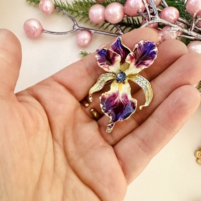 Broche em forma de flor esmaltada com cristais numa mão, fundo com ramos e bagas cor-de-rosa.