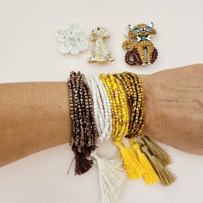 Braço com cinco pulseiras de contas coloridas e três broches decorativos sobre superfície branca.