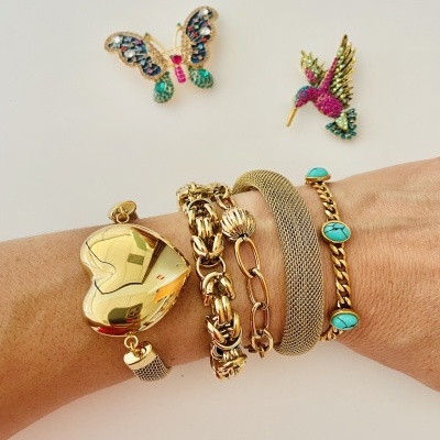 Pulso com quatro pulseiras douradas e dois broches coloridos sobre fundo branco